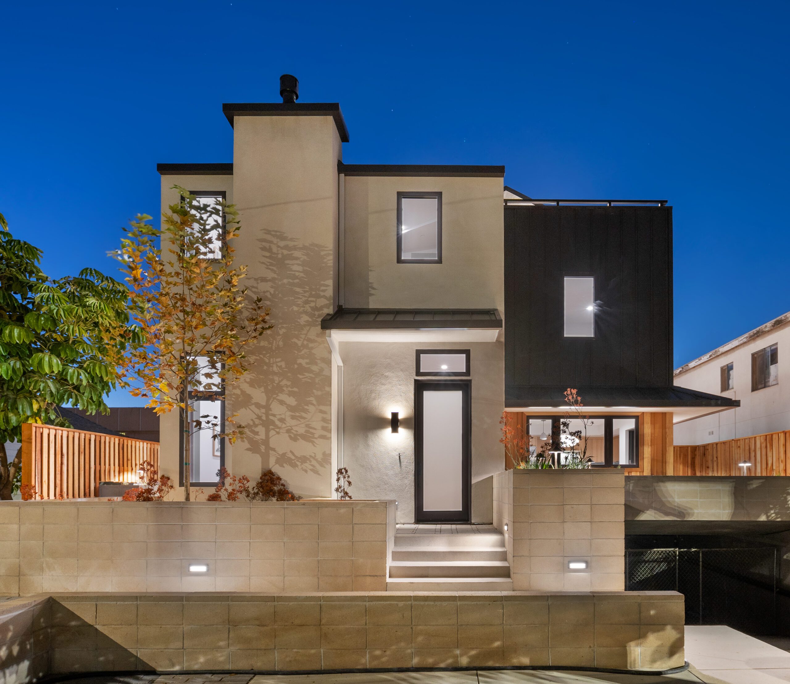 A white stucco building with arched windows and a tile roof on a street corner, surrounded by trees and parked cars on a clear day.